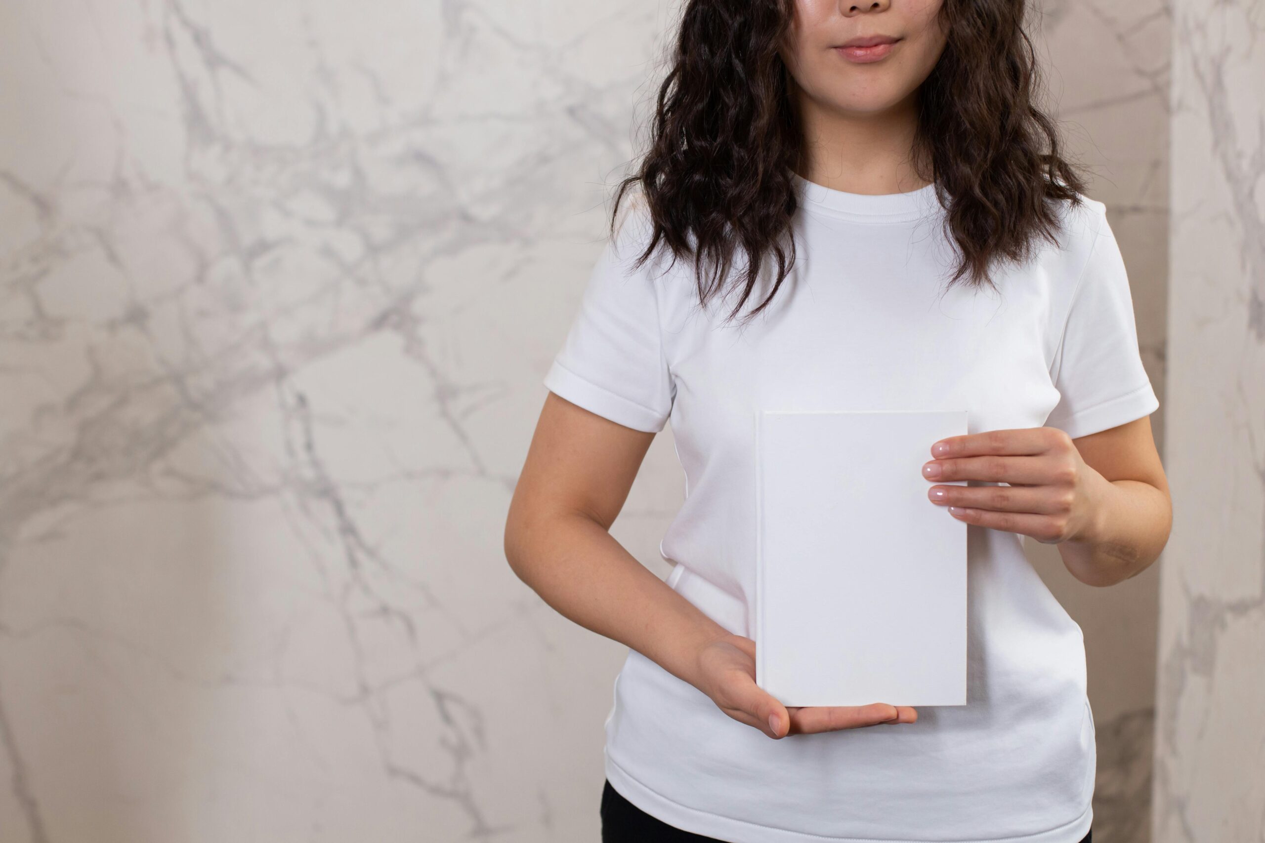 Unrecognizable female with black hair and casual t shirt showing white black notepad while standing near wall in light room
